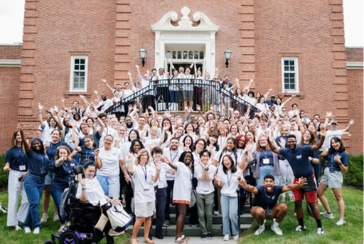 Yale Divinity School Large diverse group of college students posing with raised hands on the steps of a historic brick campus building during an outdoor university event, including participants with visible disabilities.